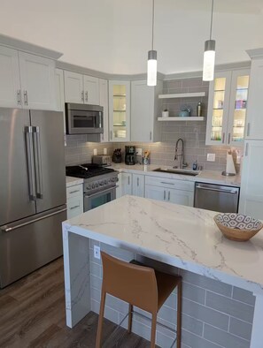 A modern kitchen with stainless steel appliances, white cabinetry, and a marble island. - DP Lantern District Townhouse STR-23-0105 (Dana Point)