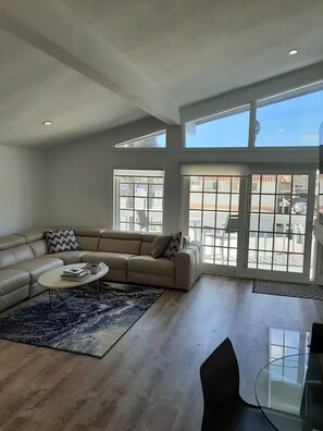 A spacious living room with a beige sectional sofa, a coffee table, and a glass dining table. Large windows provide a view of the outside. - DP Lantern District Townhouse STR-23-0105 (Dana Point)