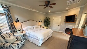 A bedroom with a bed, a chair, a small table, a plant, a fireplace, and a TV mounted on the wall. - Luxuriously Restored Early American Farmhouse (Cañon City)