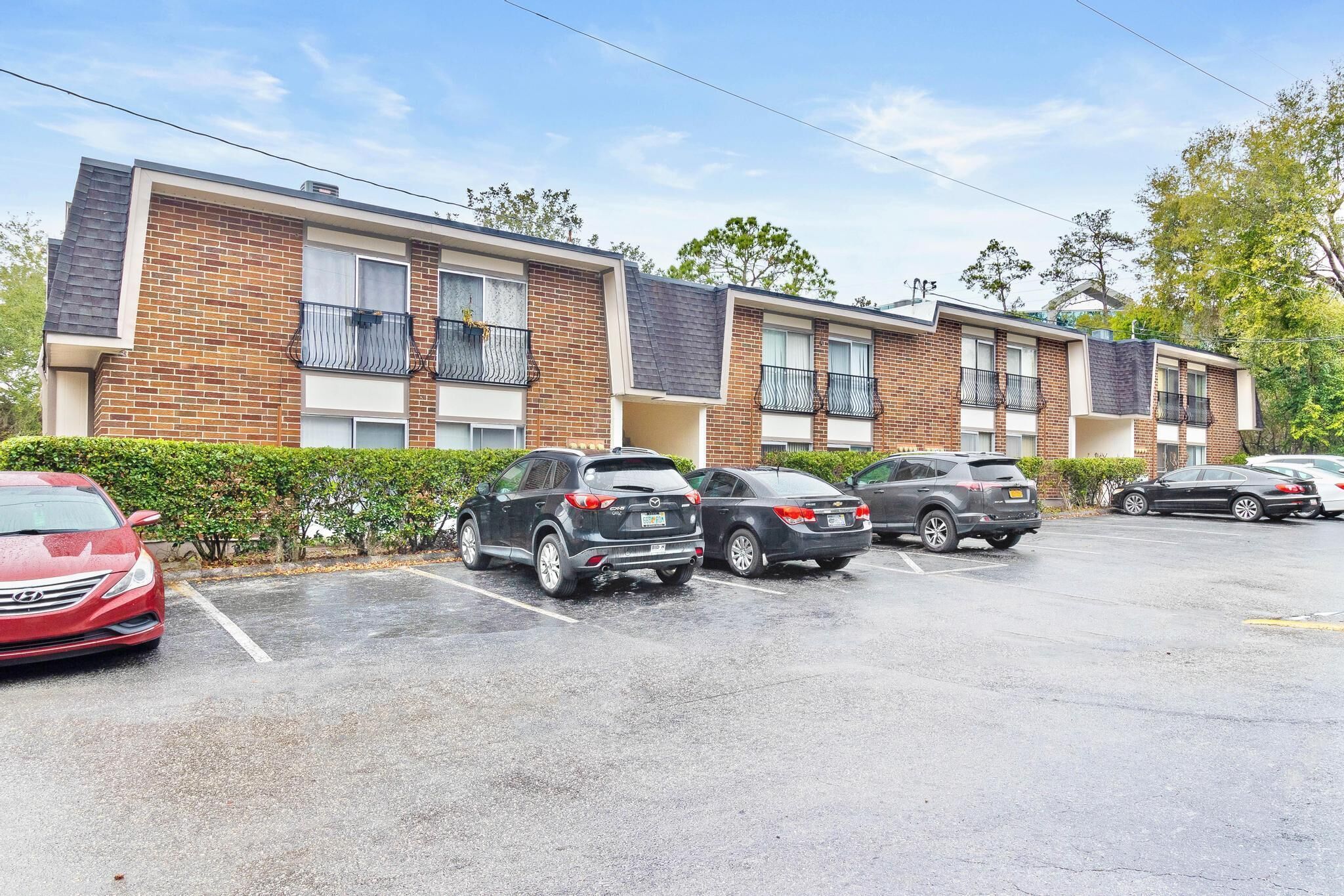 Uma fileira de edifícios de apartamentos modernos de tijolo com varandas, um estacionamento com vários carros e um céu limpo.