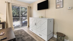 Una habitación con puerta corredera de vidrio, un armario blanco, un televisor de pantalla plana y un cuadro en la pared.