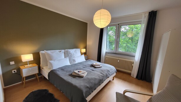 Ein modernes Schlafzimmer mit einem großen Bett, Nachttischlampen, einer Heizung und Blick auf Grün hinter dem Fenster.