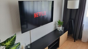 Ein modernes Wohnzimmer mit einem Wand-Fernseher, der "NETFLIX" anzeigt, einer kleinen Topfpflanze, einem Glaskerzenhalter und einer Lampe auf einer Holzkonsole.