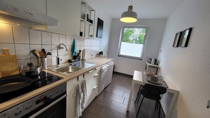 Eine moderne Küche mit weißen Schränken, einer Edelstahl-Spüle und einem schwarzen Herd. Es gibt ein Fenster mit Jalousien, einen kleinen Esstisch mit Stühlen und eine Pendelleuchte.