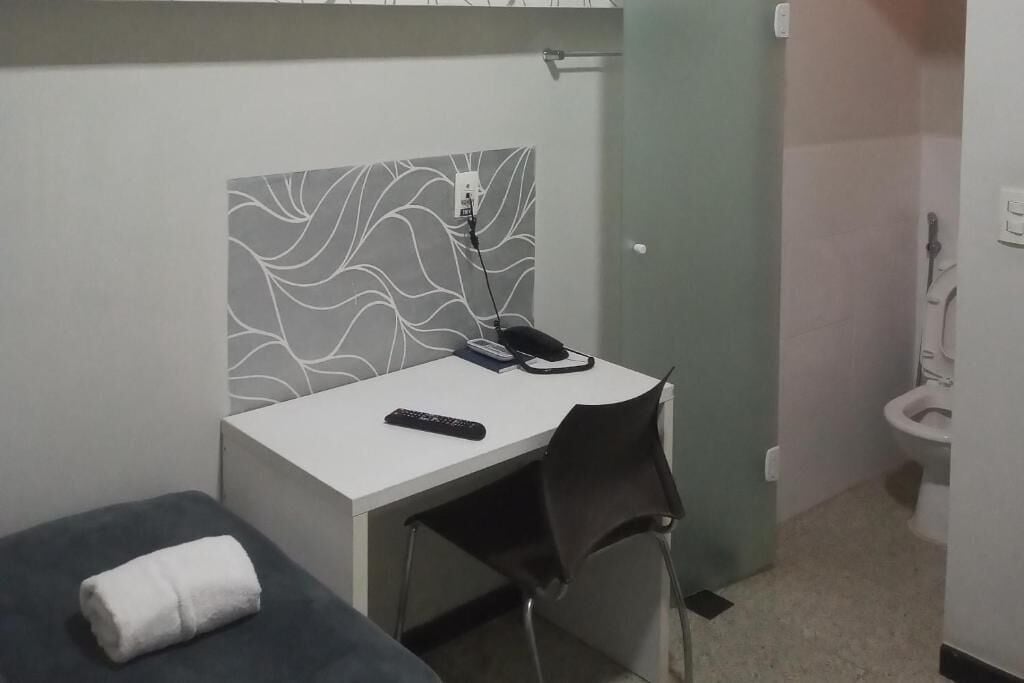 Una habitación pequeña y privada con escritorio, silla y una cama individual con una toalla enrollada.