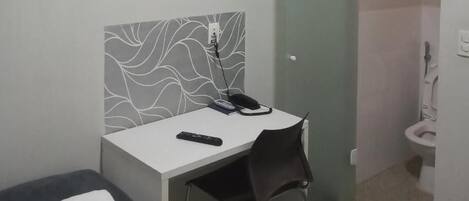Una habitación pequeña y privada con escritorio, silla y una cama individual con una toalla enrollada.