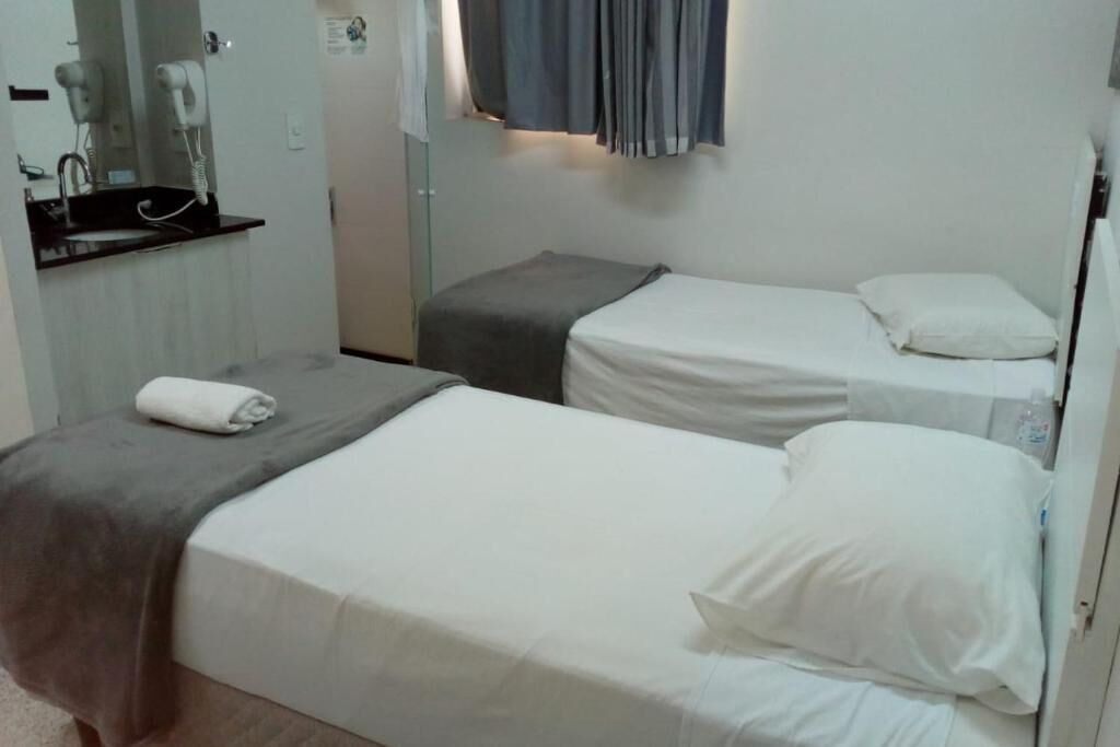 Habitación de hotel con dos camas individuales, una mesita de noche pequeña, un espejo y una ventana con cortinas.