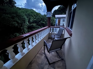 A balcony with two chairs, a white balustrade, and a view of greenery and a building in the background. - Adorable 3-bedroom apartment with AC in Elms Hall Roseau. (Elms Hall, Roseau)