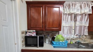 A kitchen with wooden cabinets, a microwave, a sink, and a window with lace curtains. - Adorable 3-bedroom apartment with AC in Elms Hall Roseau. (Elms Hall, Roseau)