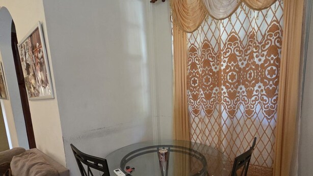 A dining area with a glass table and four chairs, a sofa, and a decorative curtain. - Adorable 3-bedroom apartment with AC in Elms Hall Roseau. (Elms Hall, Roseau)