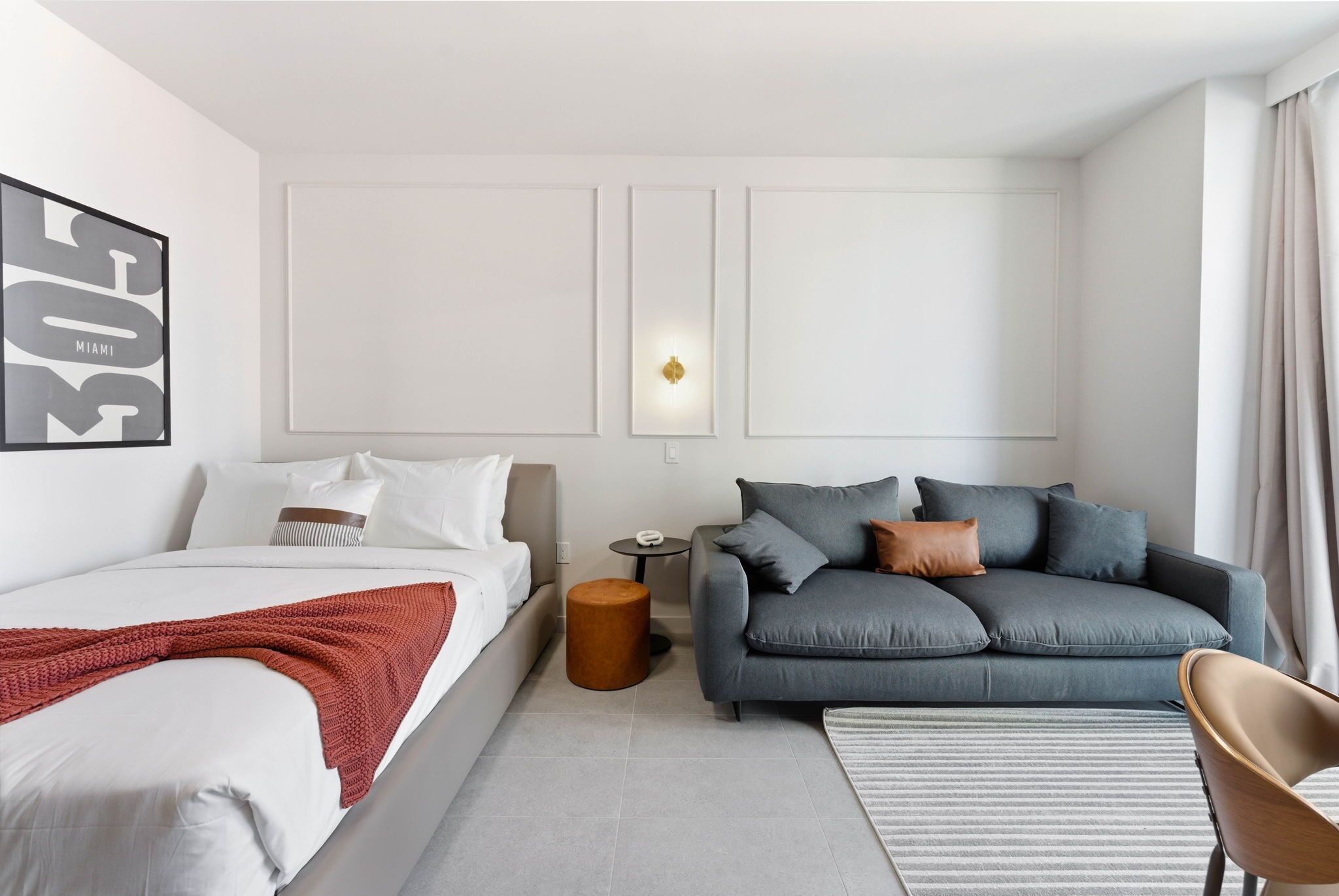 Ett modernt hotellrum med en säng, en grå soffa, ett litet runt bord och en väggmonterad lampa.