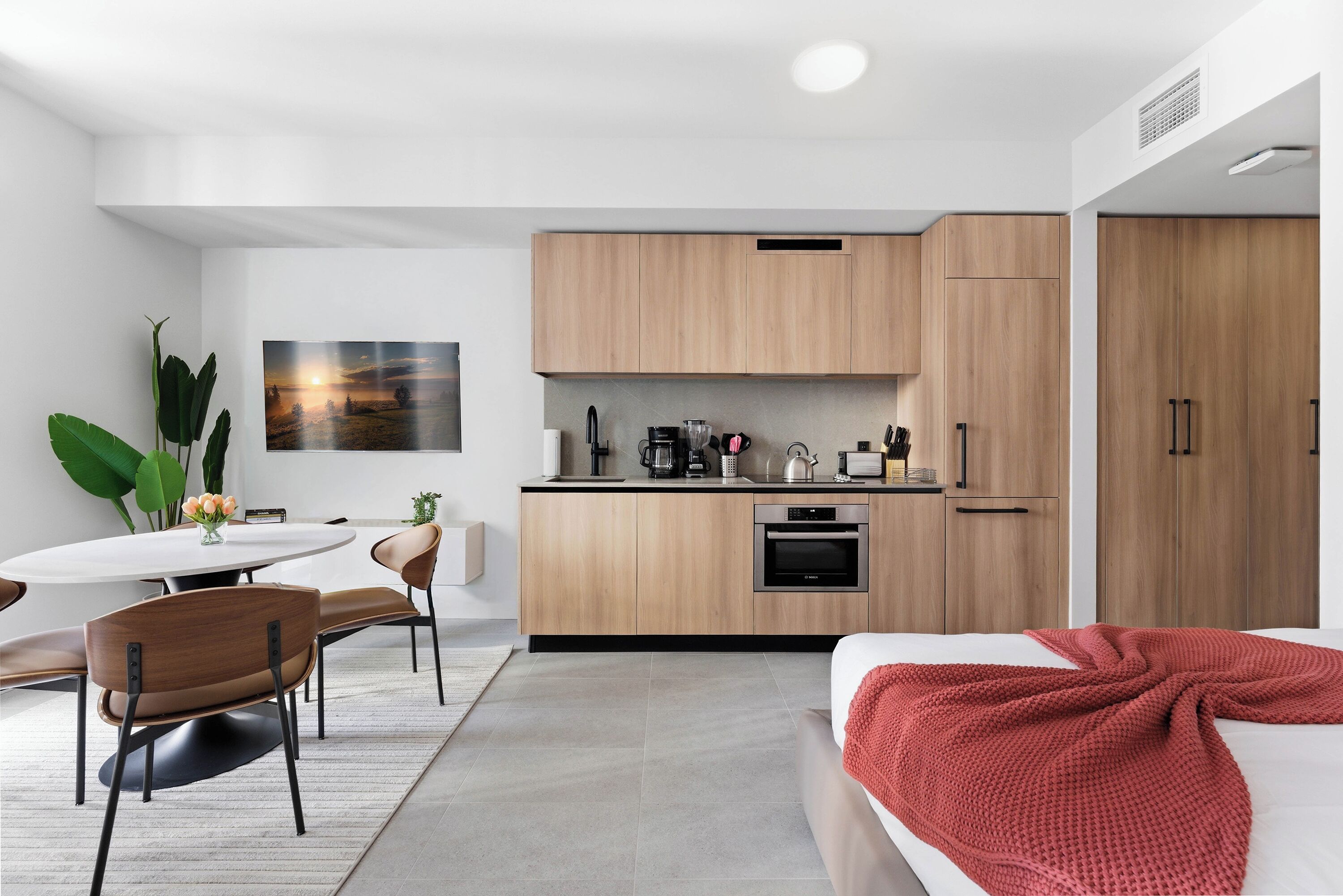 A modern, compact hotel room with a kitchenette, a dining area with a round table and chairs, and a bed with a red blanket.