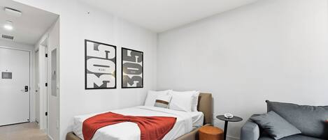 A modern bedroom with a bed, a grey sofa, a small round table, and two wall-mounted artwork pieces.