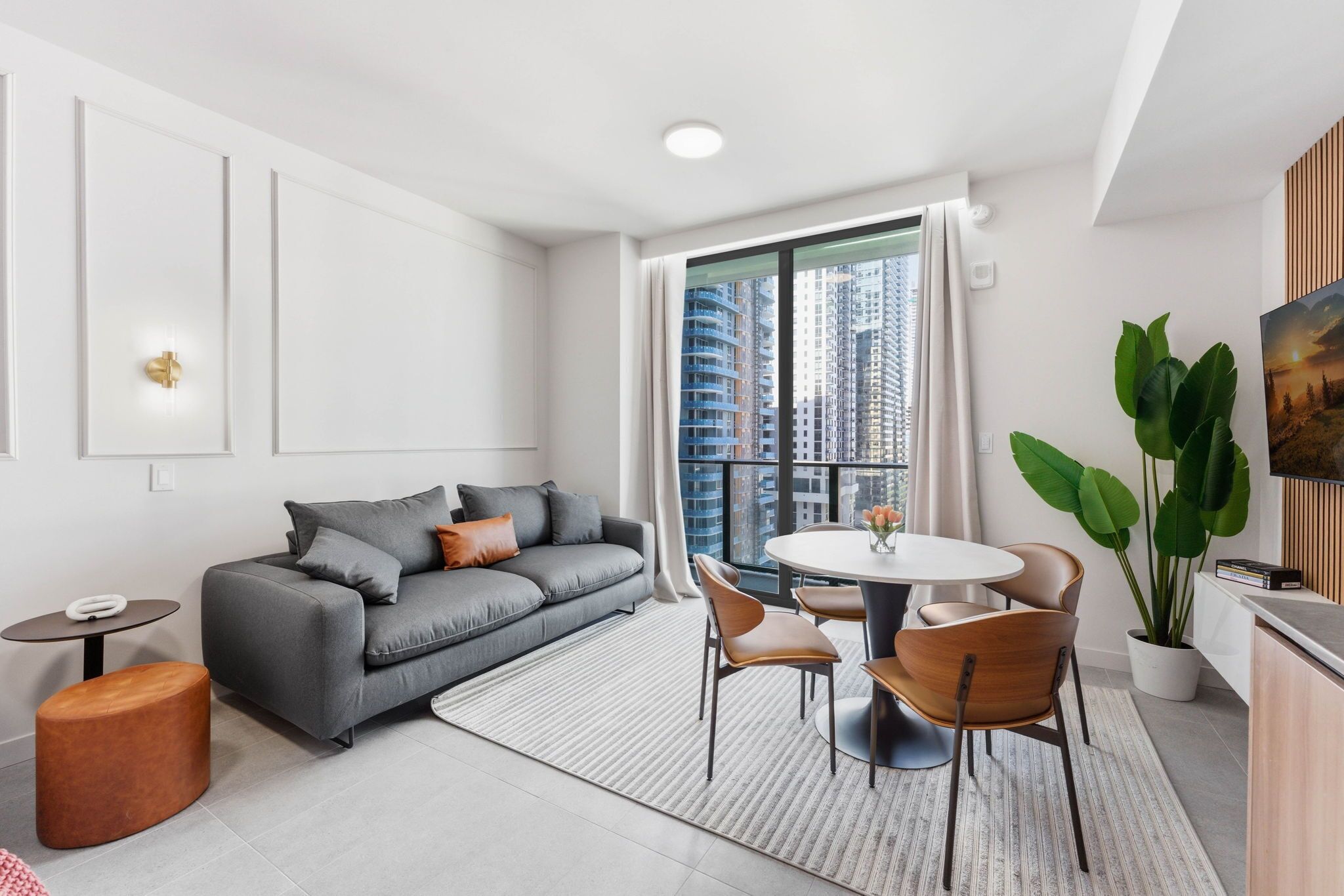 A modern living room with a grey sofa, a round dining table with chairs, a small side table, a potted plant, and a view of tall buildings through a large window.