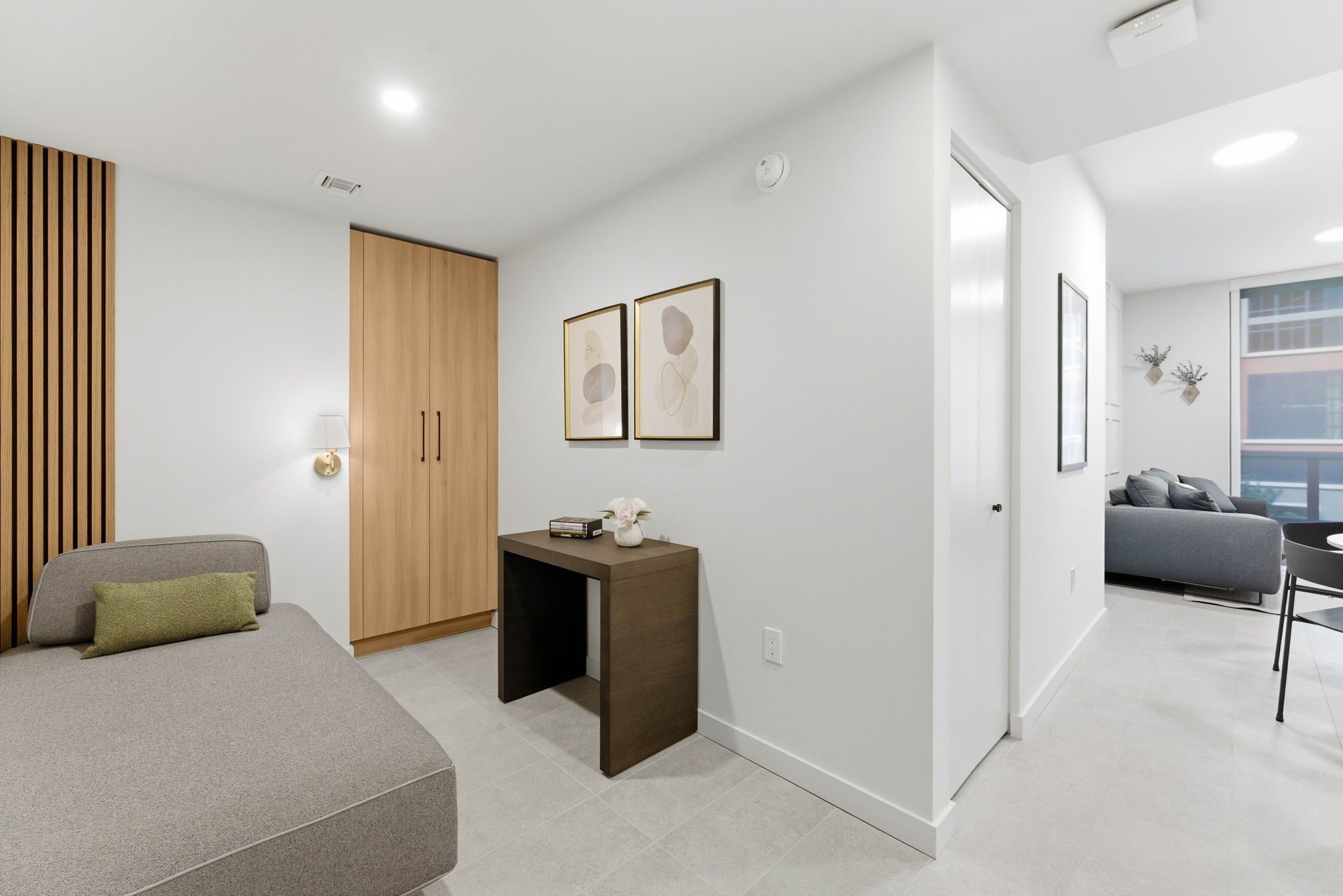A modern living room with a sofa, a small table with a vase, and a wardrobe. There are framed pictures on the wall and a grey couch in the background.