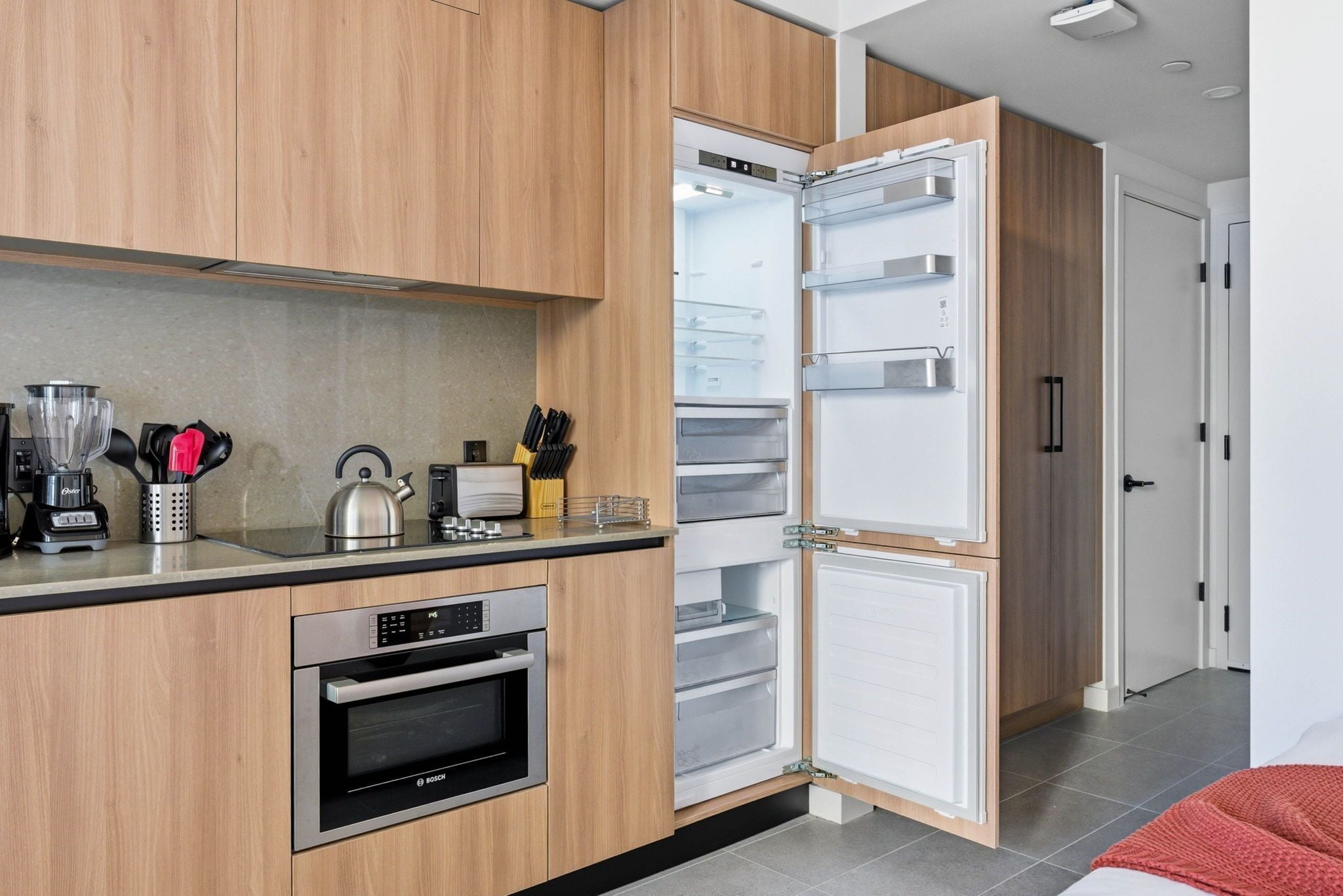 Une cuisine moderne avec des meubles en bois, un four Bosch, un réfrigérateur encastré et une bouilloire posée sur le plan de travail.