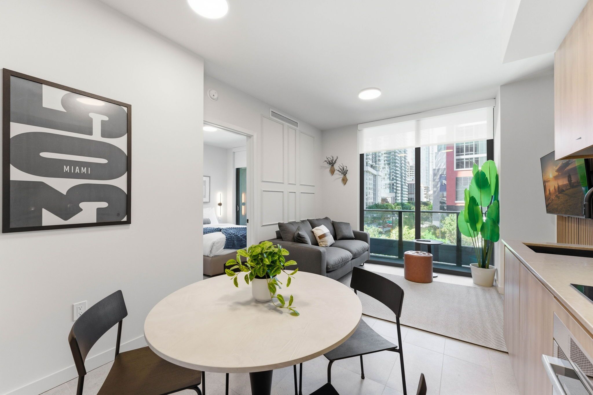 Een moderne woonkamer met een grijze bank, een ronde eettafel met stoelen, een potplant en een groot raam met uitzicht over de stad.
