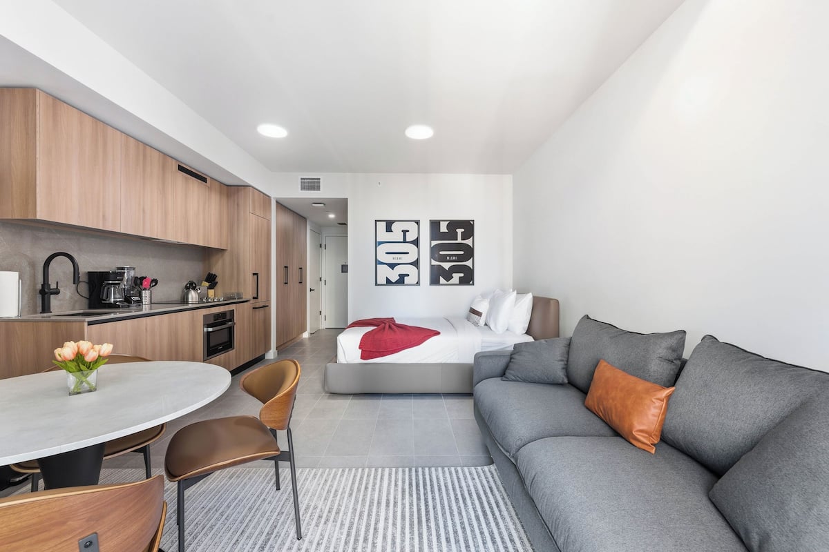 A modern hotel room with a kitchenette, a dining area, and a large gray sofa with a red throw blanket.