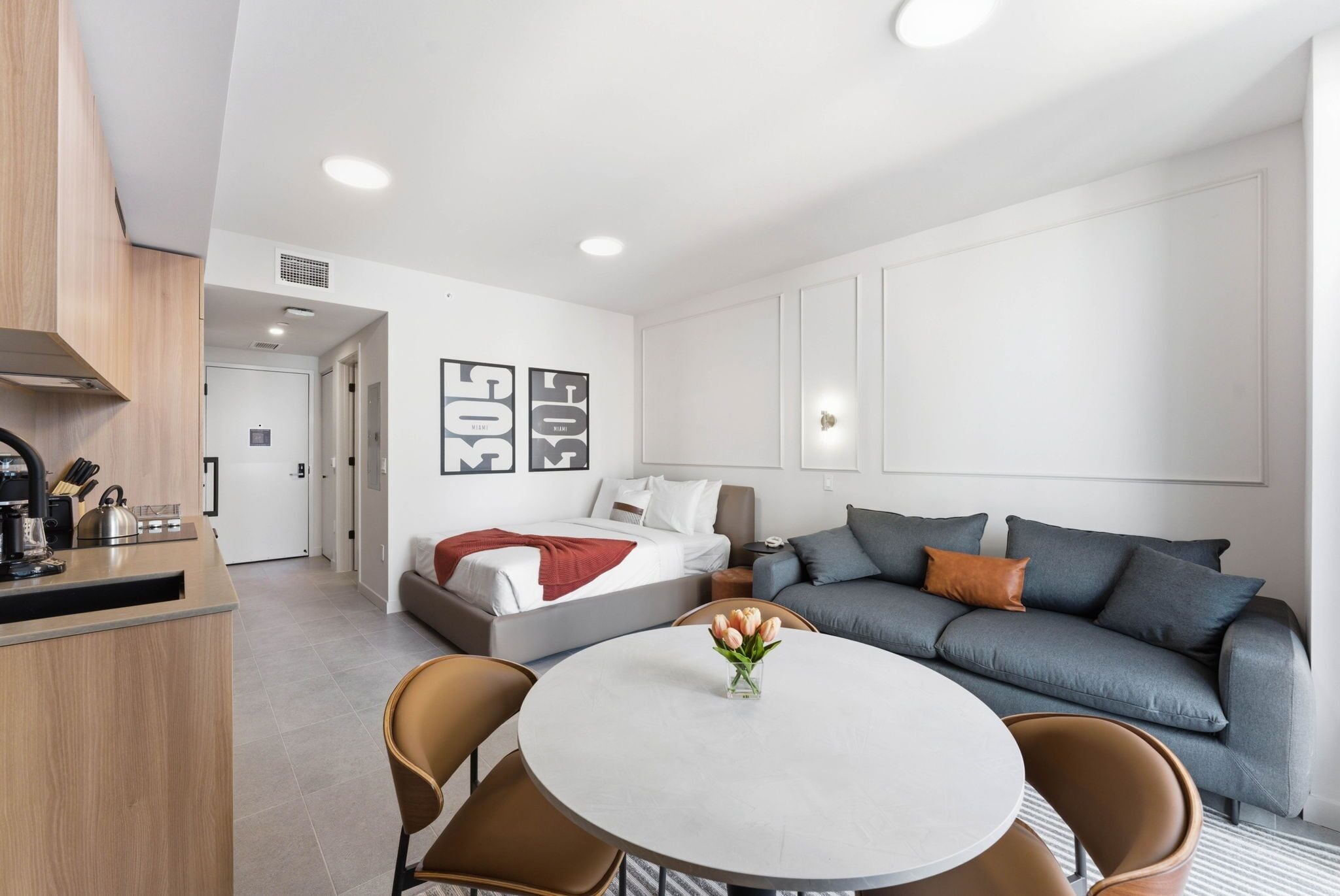 A modern hotel room with a kitchenette, a bed with a red blanket, a grey sofa, and a round dining table with chairs.