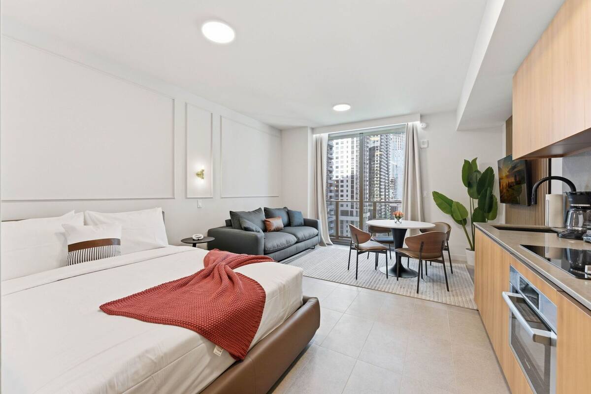 A modern hotel room with a large bed, a sofa, a dining area, and a kitchenette.