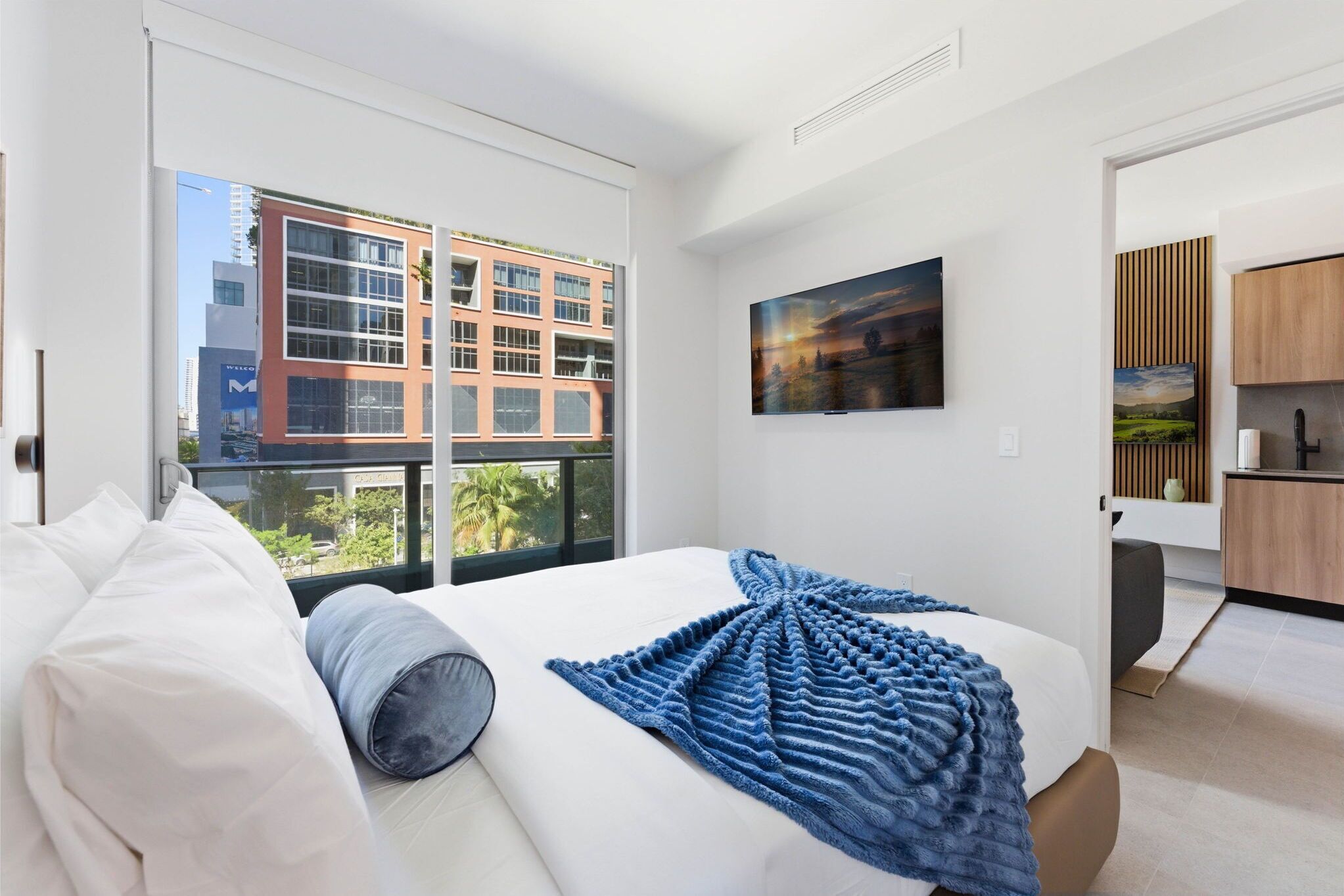 A modern hotel room with a large bed, a view of a building outside, and a kitchen area visible through an open door.