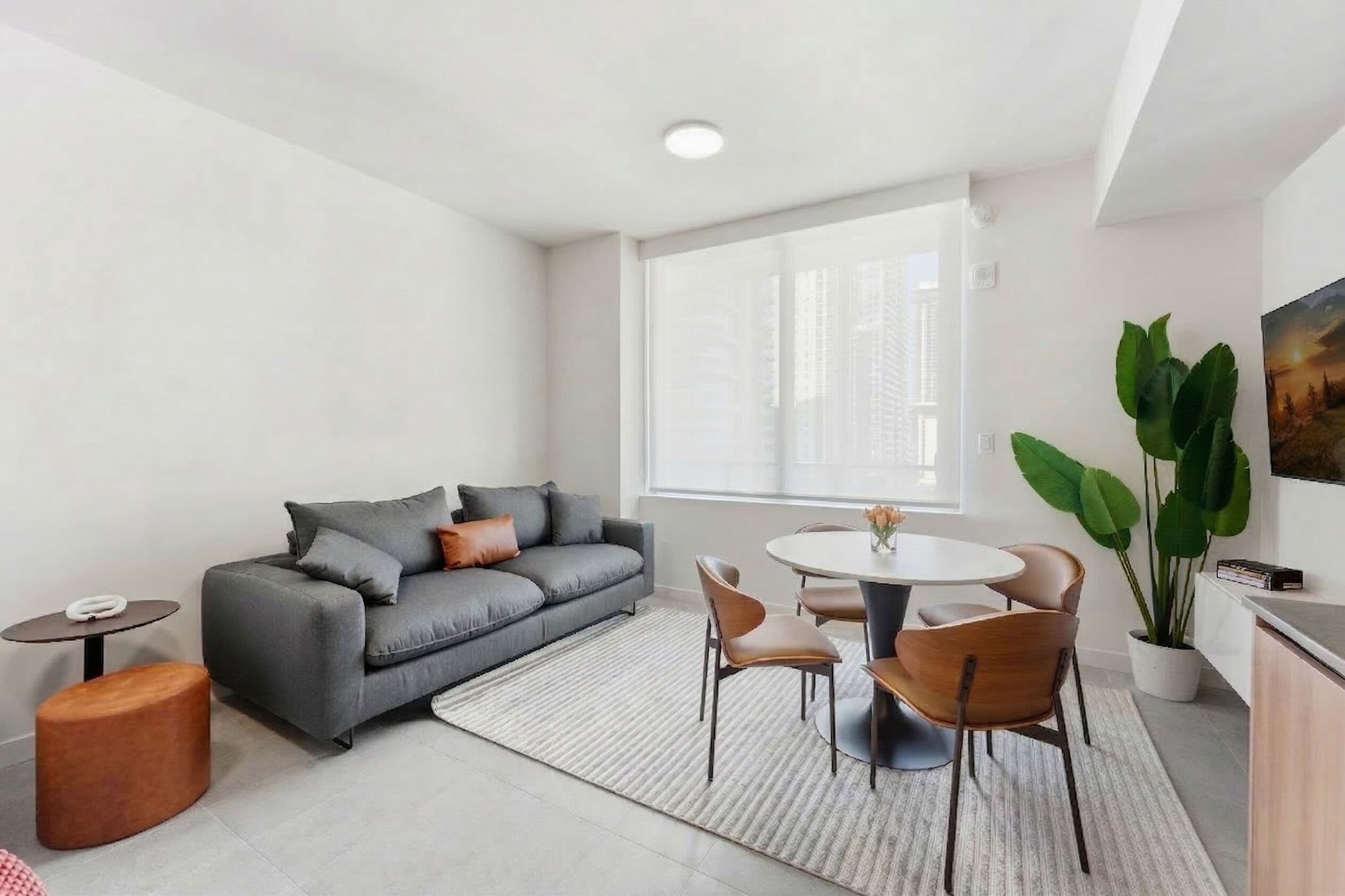 A modern living room with a grey sofa, round dining table, and chairs. There is a window with blinds, a potted plant, and a painting on the wall.