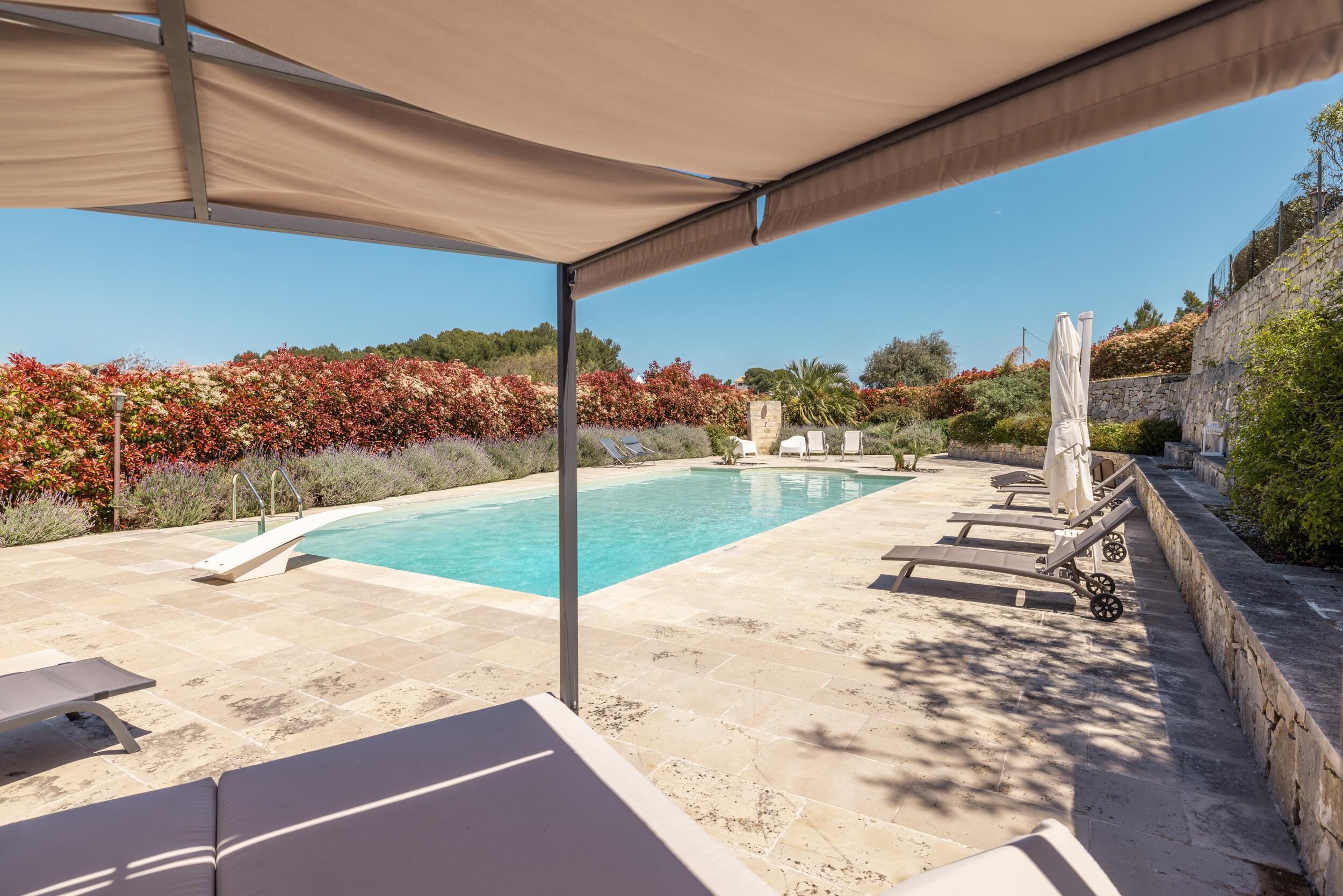 A pool area with lounge chairs and a canopy, surrounded by landscaped gardens and a stone wall.