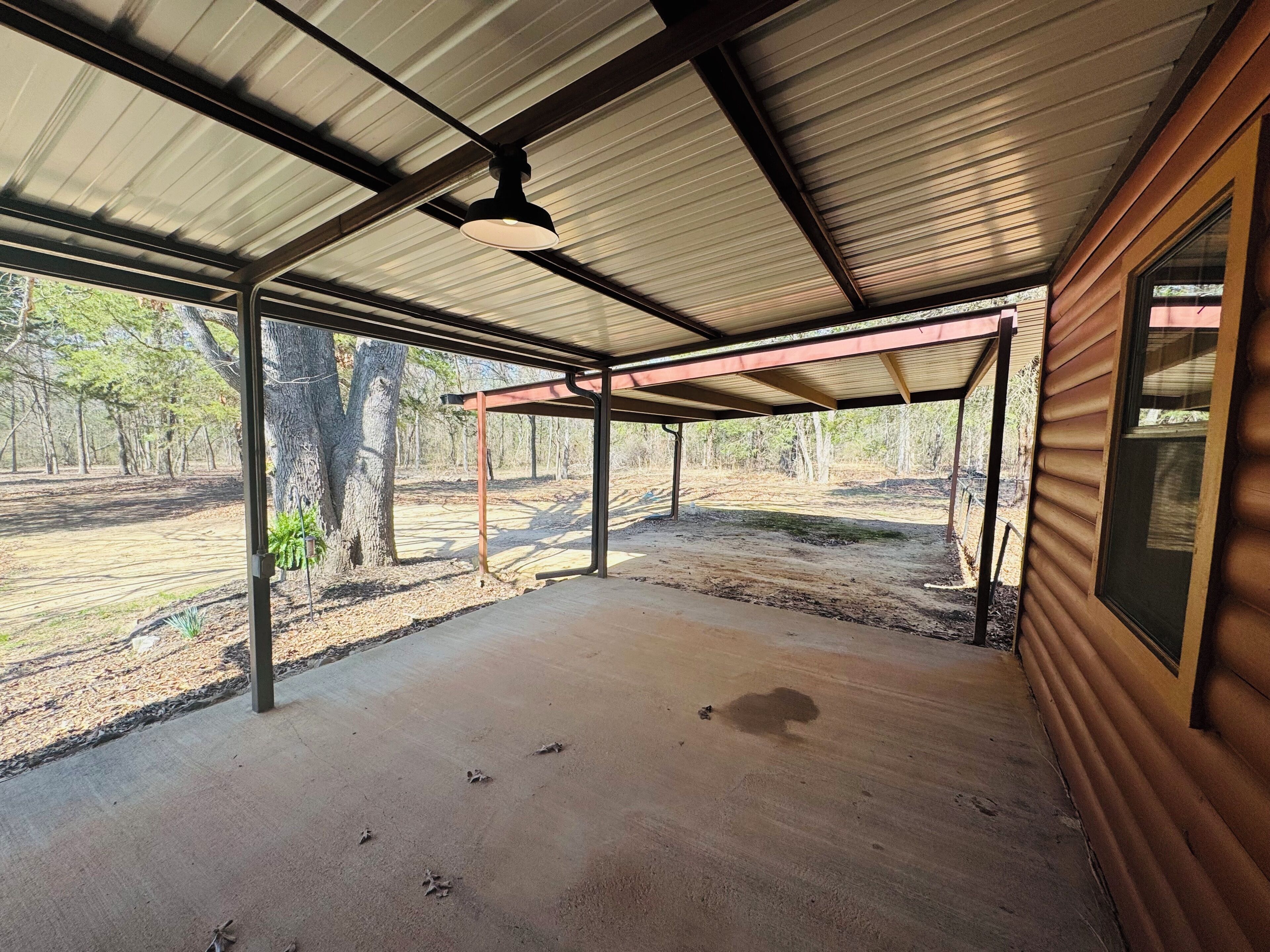 Un espace extérieur abrité, doté d’un sol en béton, de poutres métalliques et offrant une vue sur une zone boisée.