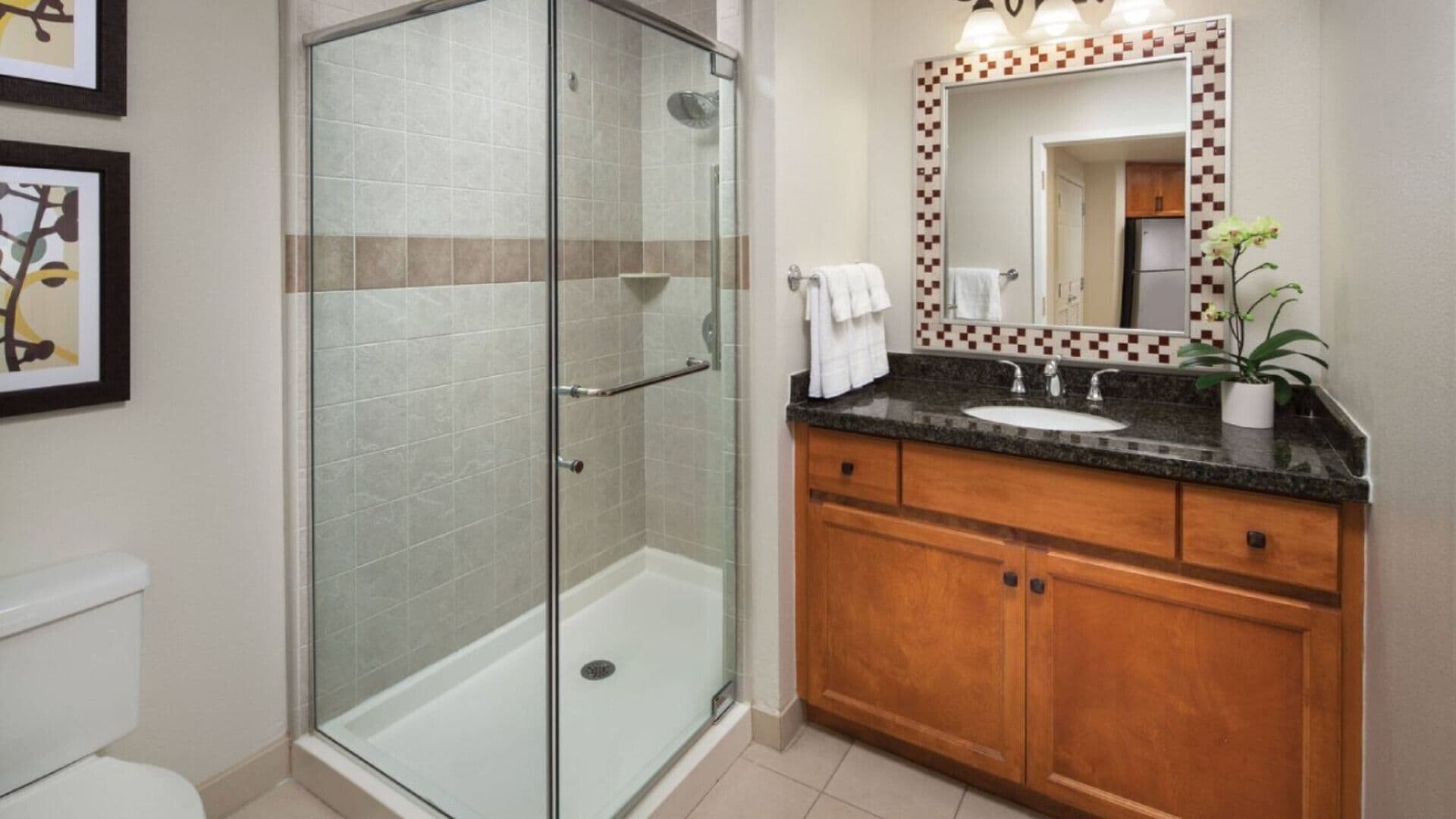 Een badkamer met een douche met glazen wand, een wastafel met houten kastje en een spiegel met decoratieve tegelrand.