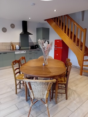 Un comedor con una mesa de madera redonda, sillas de mimbre y un florero con flores secas. Se observa un refrigerador rojo y una cocina con estufa y armarios al fondo.