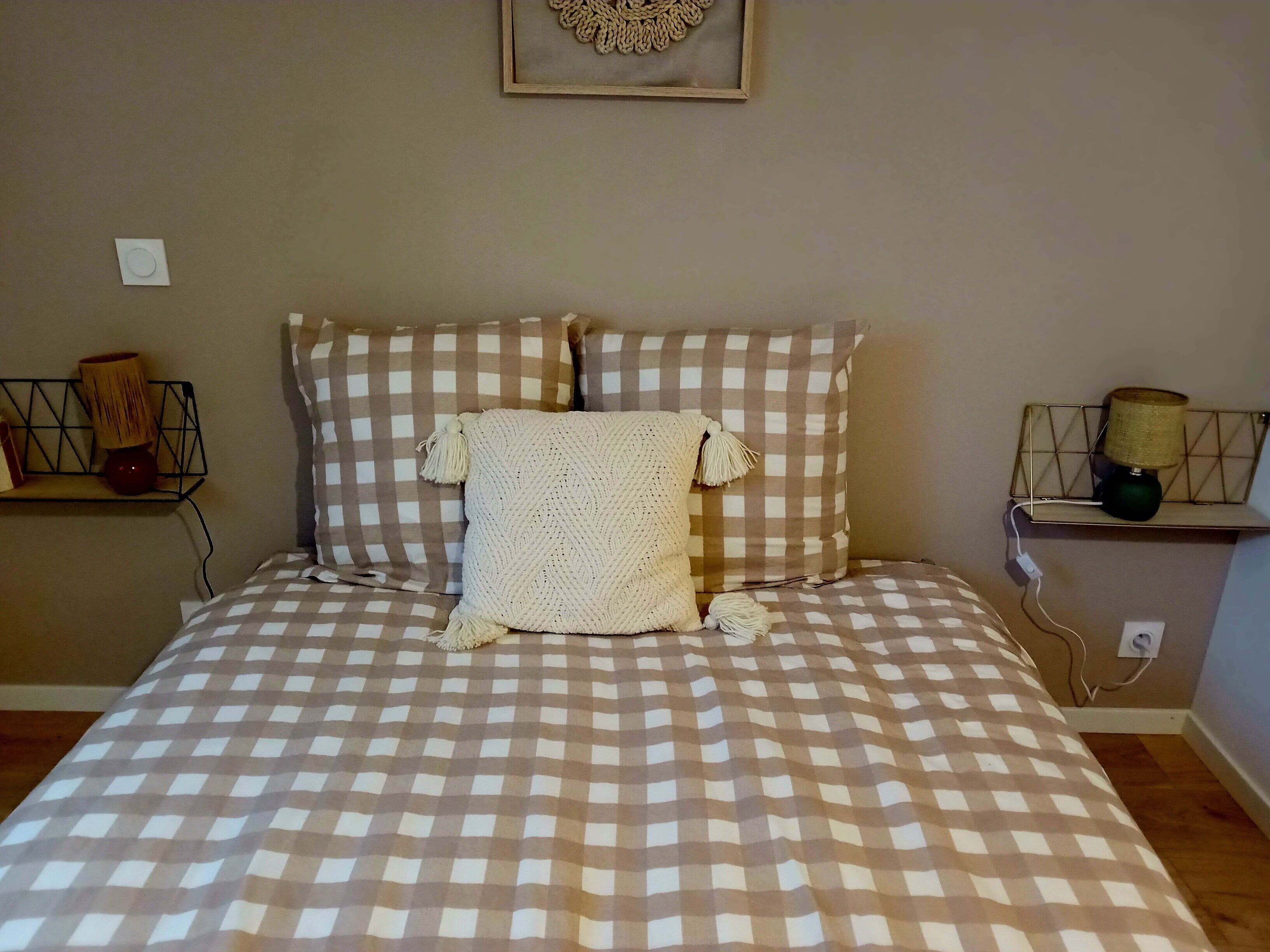 Een bed met een geruit sprei, een gebreide plaid en een metalen plank met een lamp.