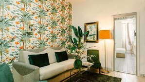A living room with a floral wallpaper, a sofa with cushions, a green armchair, a floor lamp, and a mirror on the wall. - Random Apartment Deluxe (Naples)