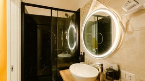 A modern bathroom with a glass shower enclosure, a round mirror with a light, and a white sink. - Random Apartment Deluxe (Naples)