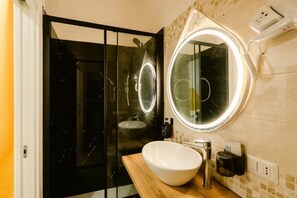 A modern bathroom with a glass shower enclosure, a round mirror with a light, and a white sink. - Random Apartment Deluxe (Naples)