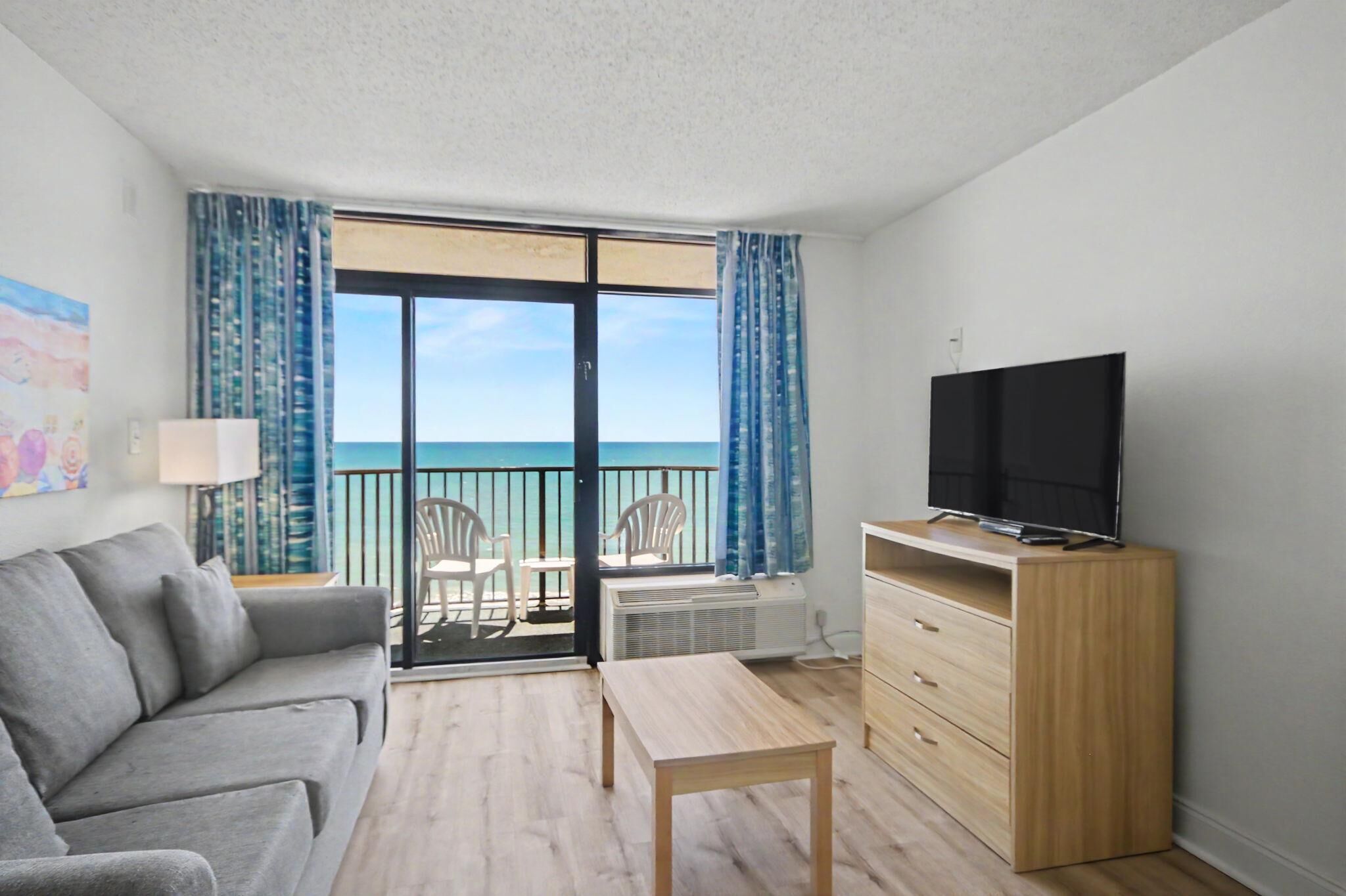 A room with a sofa, a TV on a stand, a table, a dresser, and a balcony with a view of the sea.