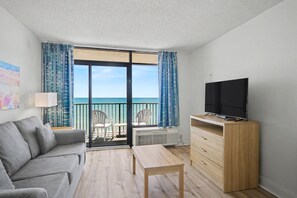 Ein Zimmer mit einer Couch, einem Fernseher auf einem Schrank, einem Tisch, einer Kommode und einem Balkon mit Meerblick.