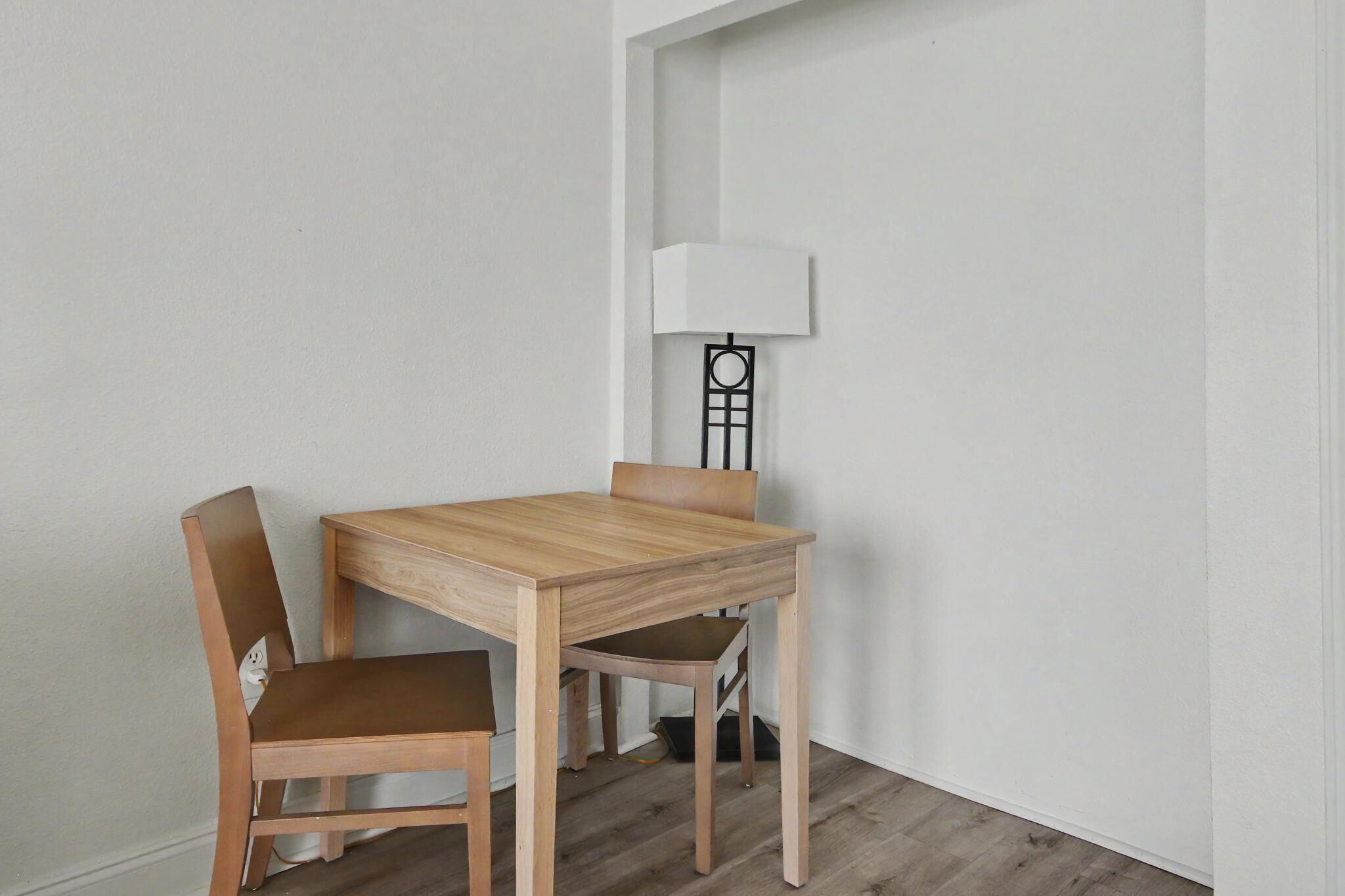 A corner room with a wooden table and two chairs, a white wall, and a floor lamp.