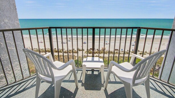 Un balcon avec deux chaises en plastique blanches et une petite table, donnant sur une plage où l’on peut voir des gens et des véhicules.