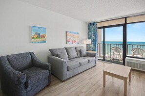 Une pièce avec un canapé gris, un fauteuil gris, une table en bois et une vue sur l’océan par de grandes fenêtres.