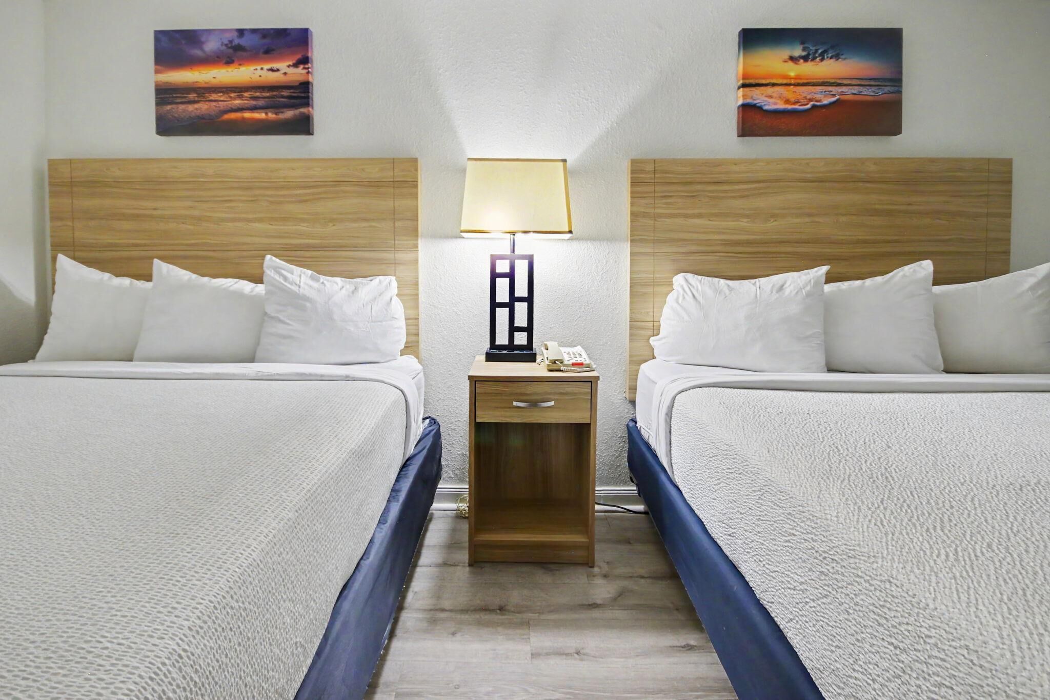 Hotellihuone, jossa on kaksi sänkyä, yöpöytä lampun kanssa ja kaksi taulua seinällä.