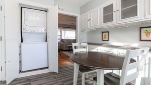 Une cuisine compacte dotée d’un lave-linge et d’un sèche-linge intégrés, un coin repas avec une table en bois et des chaises blanches, et un espace de vie avec un canapé et une table basse.