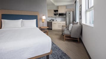 Hotellihuone, jossa on sänky, pieni pöytä, tuoli, pieni keittiönurkkaus ja ikkuna, jossa on verhot.