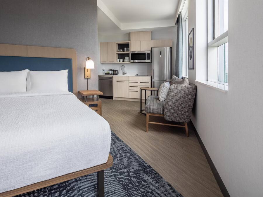 Hotellihuone, jossa on sänky, pieni pöytä, tuoli, pieni keittiönurkkaus ja ikkuna, jossa on verhot.