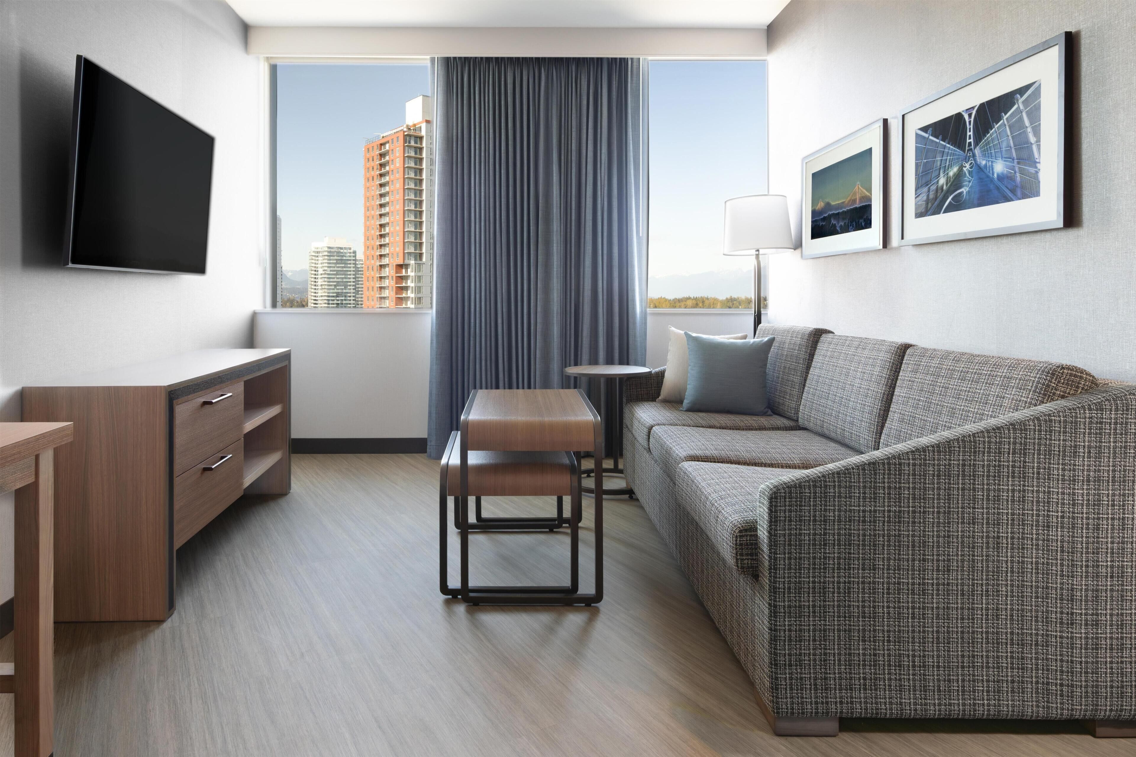 A modern hotel room with a flat-screen TV, a sofa, a small table, a desk, and a view of a cityscape through a large window.