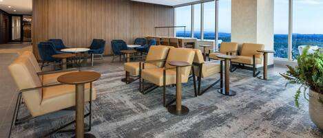 A modern lounge with leather chairs, round tables, and large windows offering a city view.