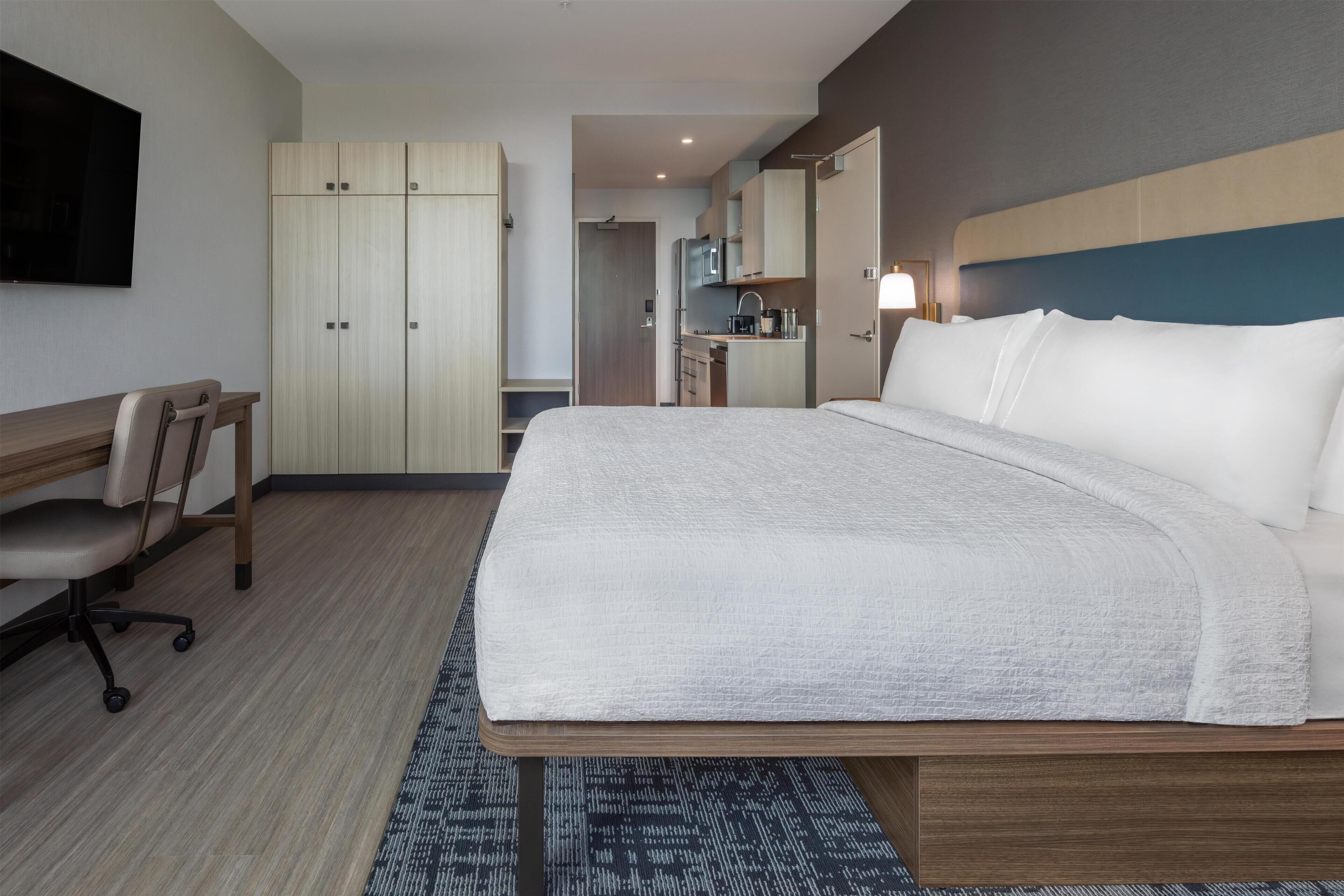 Habitación de hotel con cama, escritorio, silla, armario, kitchenette y televisión.
