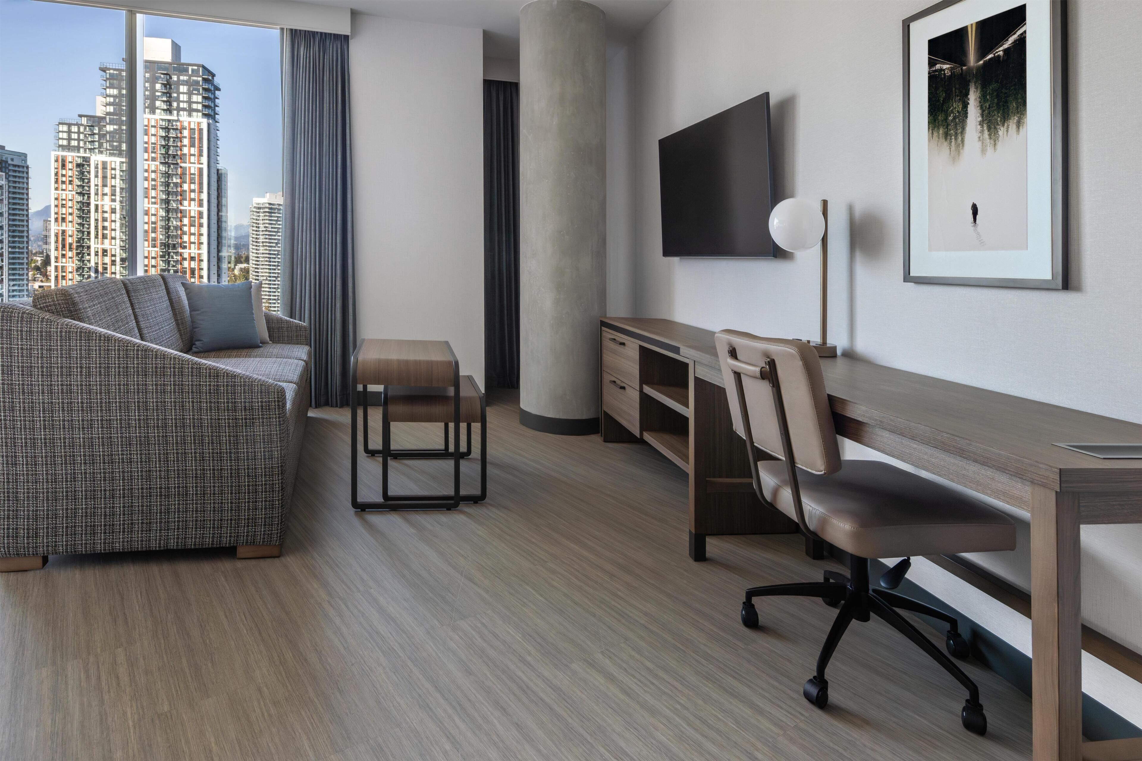 A modern hotel room with a sofa, desk, chair, TV, and a large window with a city view.