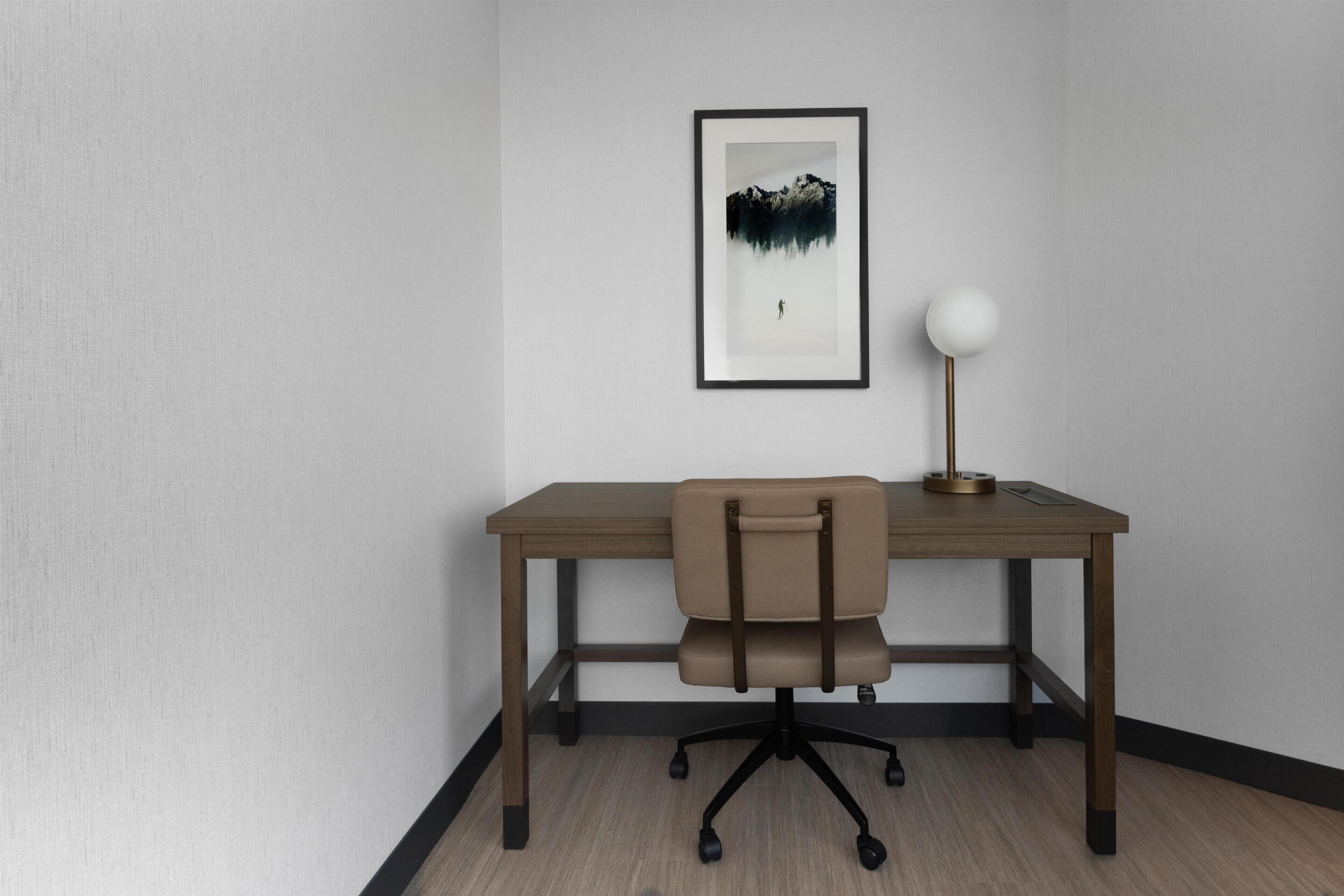 A room with a desk, chair, framed artwork, and a lamp.