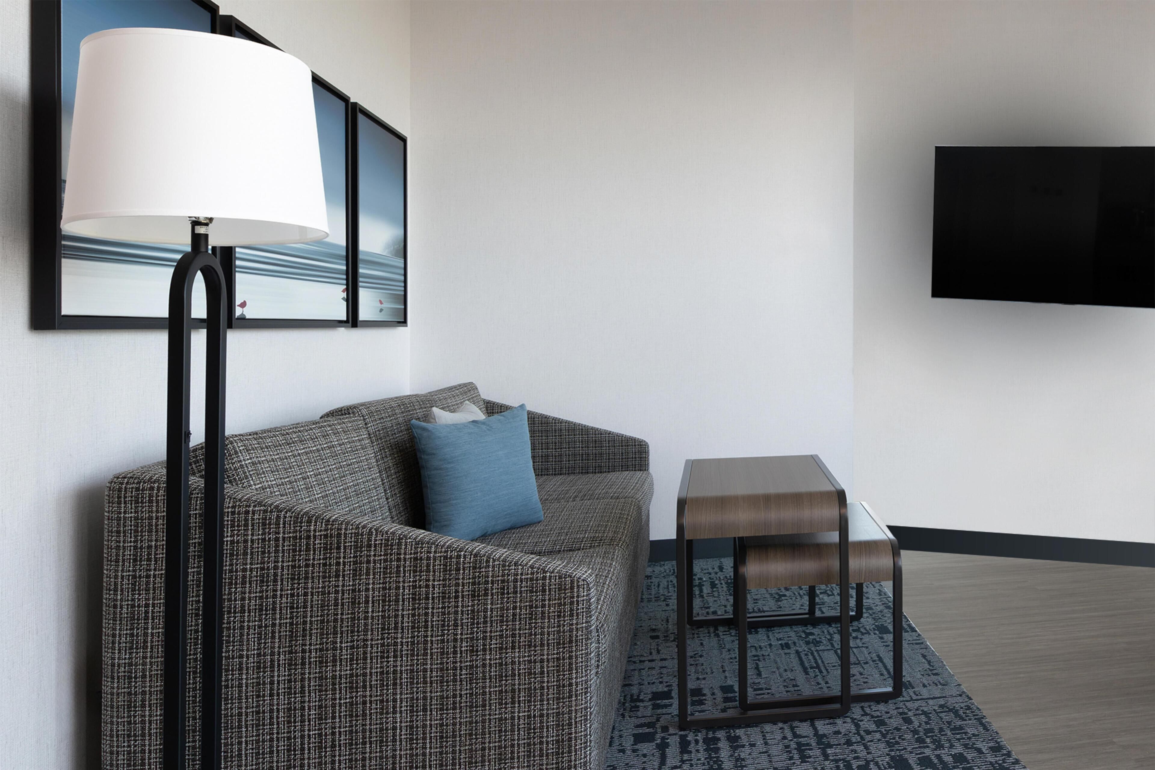 Ein modernes Hotelzimmer mit einer Couch, einem Beistelltisch, einer Lampe und einem an der Wand befestigten Flachbildfernseher.