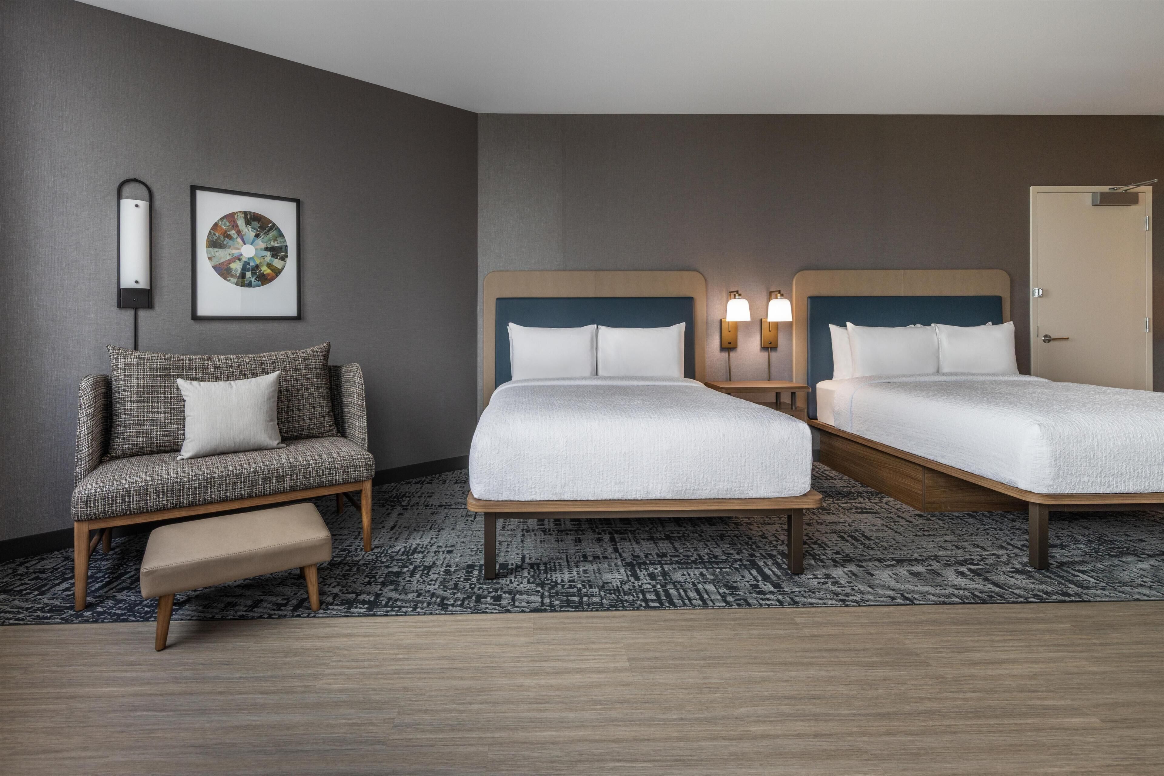 A hotel room with two beds, a sofa, a bench, a wall-mounted lamp, and a framed picture.